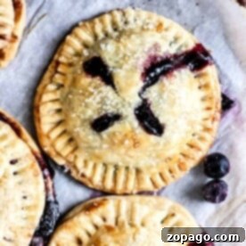 Air-Fried Blueberry Hand Pies 7 Golden-crusted air fryer blueberry hand pies, topped with sugar crystals and bursting with luscious blueberry filling, rest elegantly on a parchment-lined surface.