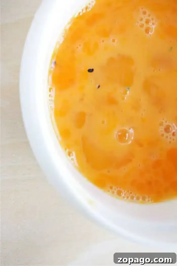 A close-up view of a bowl filled with an orange liquid, likely part of an egg wash recipe. The liquid appears smooth with a few small bubbles and specks on its surface. The bowl is white and sits on a light wooden surface.