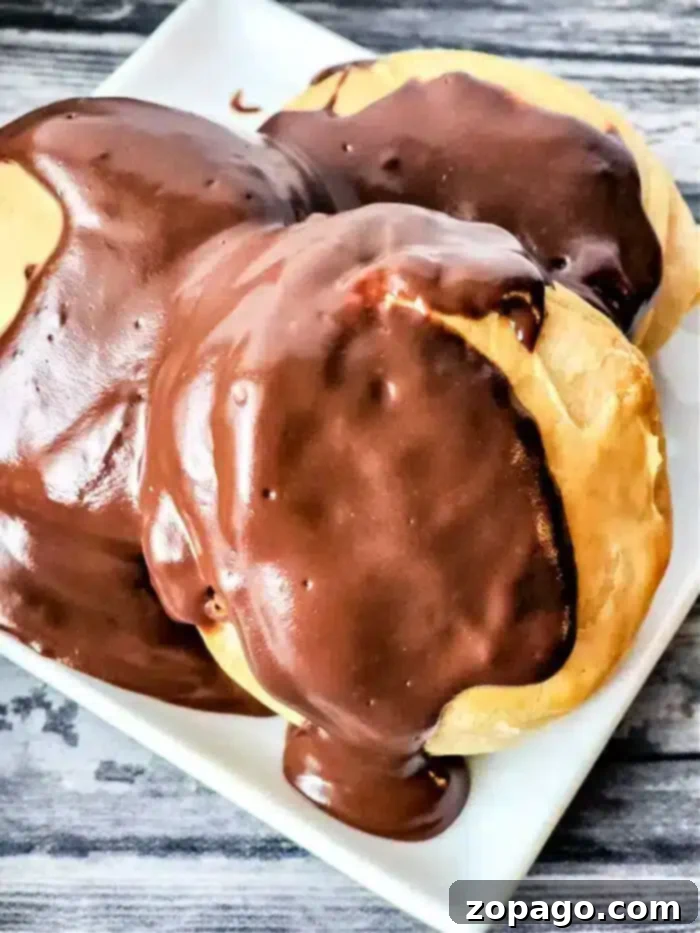 Chocolate Drenched Biscuit Dreams 2 Three cream puffs covered in thick chocolate glaze are displayed on a white rectangular plate, showcasing the richness akin to mastering how to make chocolate gravy. The background consists of a wooden surface with a gray and black grain pattern.