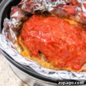The crockpot meatloaf recipe features a savory meatloaf covered in rich tomato sauce, slowly cooking to perfection in a foil-lined slow cooker.