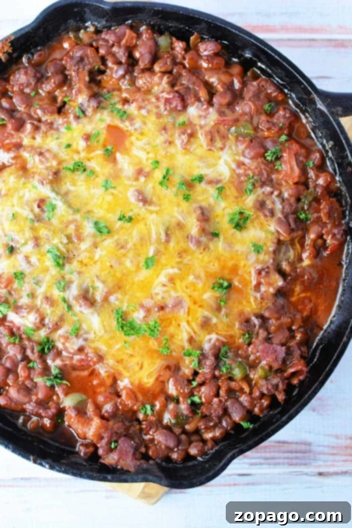 Savory Bean Medley 2 A cast iron skillet filled with baked bean casserole topped with a layer of melted cheese and garnished with chopped parsley. The dish is placed on a light wooden surface, ready to be served.