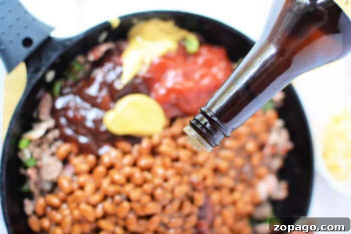 Savory Bean Medley 5 A close-up of a skillet filled with baked bean casserole, ground meat, yellow mustard, and ketchup. A bottle is seen from the top right corner, pouring additional sauce onto the mixture. The skillet handle is visible on the left side of the image.