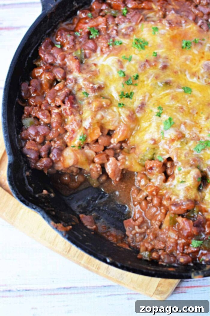 Savory Bean Medley 7 A cast-iron skillet filled with baked bean casserole topped with melted cheese and garnished with chopped parsley sits on a wooden trivet. The dish appears to be freshly cooked, with a portion already served from the skillet, showcasing its delicious appeal.