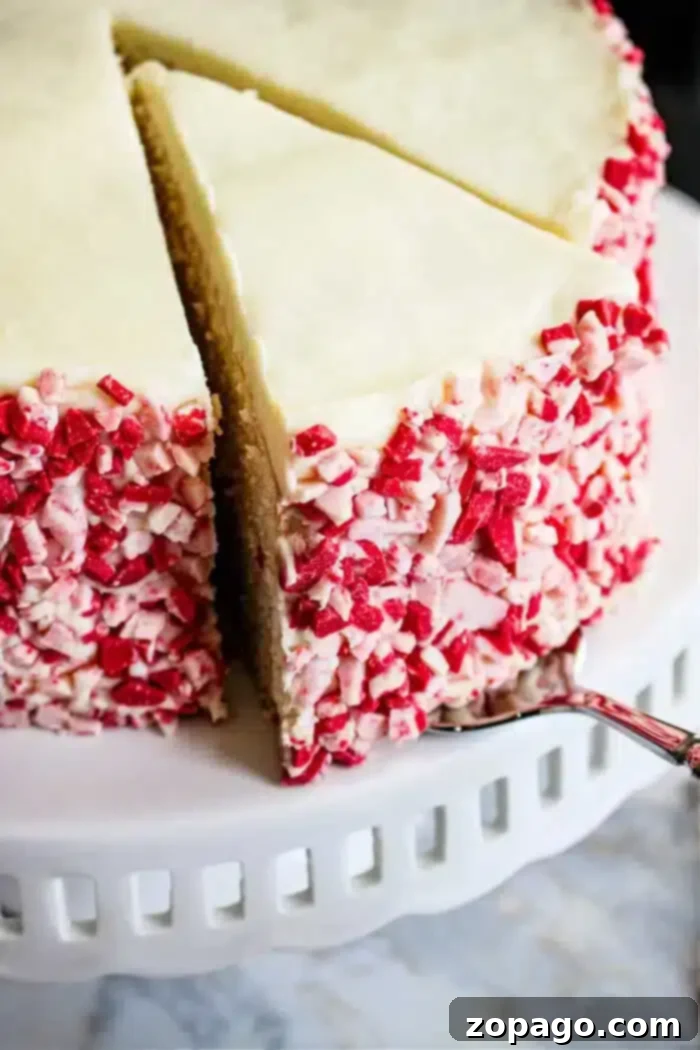 Ultimate Peppermint Bliss Cake 2 A beautifully decorated Peppermint Cake with white frosting and red and white crushed candy canes on the sides, displayed on a pristine white cake stand. A slice has been removed, revealing the moist interior.