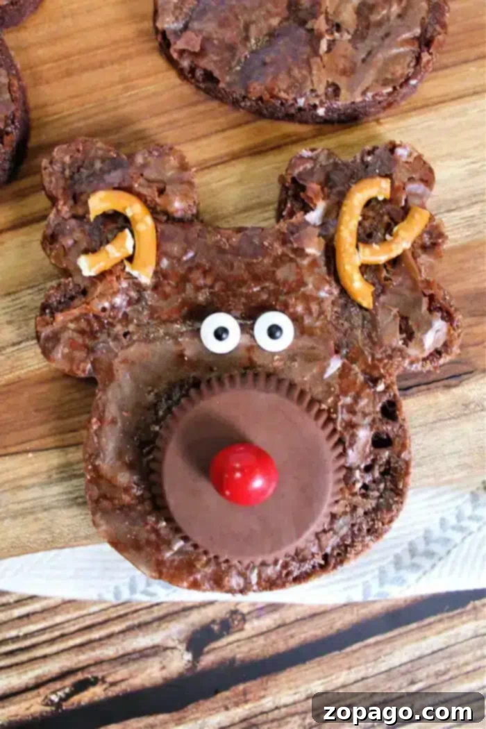Santa's Favorite Brownies 2 A reindeer-shaped Christmas brownie with a peanut butter cup for a nose, a red candy on top, candy eyes, and broken pretzels as antlers rests on a wooden surface. Other similar brownies are partially visible in the background.