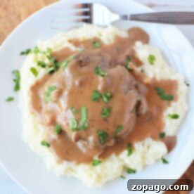 Plate of mashed potatoes topped with brown gravy and chopped herbs, served alongside tender Crockpot Salisbury Steak with a fork.