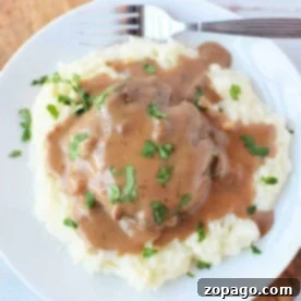 Plate of mashed potatoes topped with brown gravy and chopped herbs, served alongside tender Crockpot Salisbury Steak with a fork.