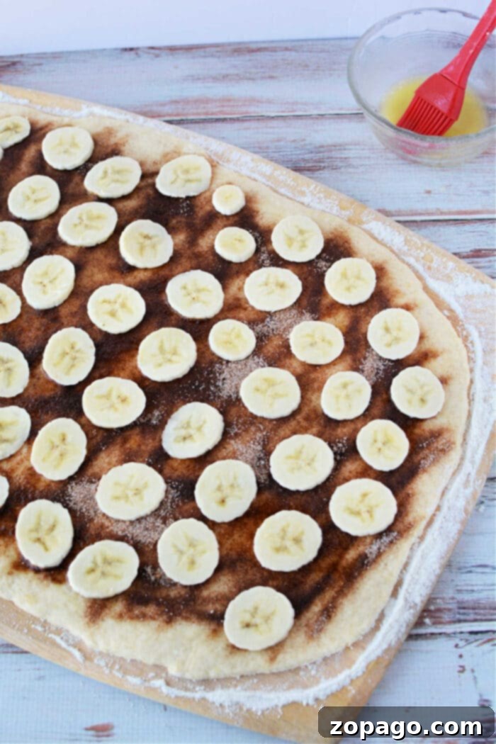 Gooey Banana Bread Cinnamon Buns 4 A wooden surface cradles cinnamon roll dough topped with a blend reminiscent of banana bread cinnamon rolls, featuring cinnamon, sugar, and sliced bananas.
