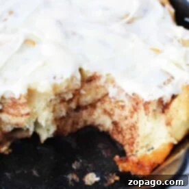 Cinnamon Swirled Banana Bread Buns 6 A close-up of banana bread cinnamon rolls with cream cheese frosting in a black skillet, one roll missing.