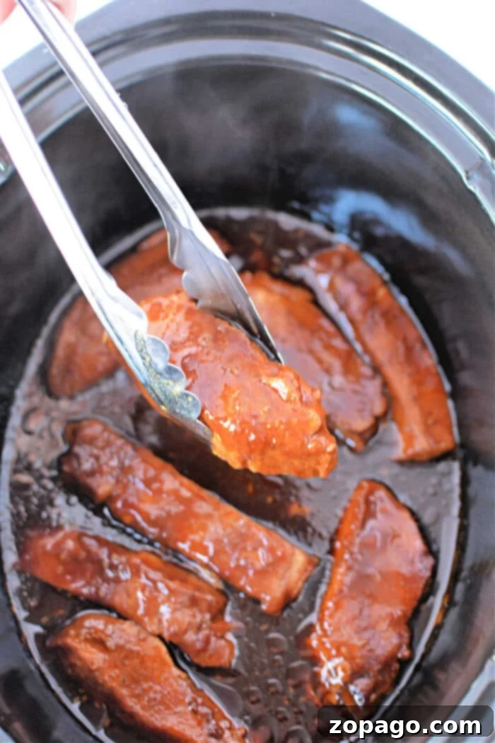 Fall Off The Bone Slow Cooker Country Ribs 7 Tongs holding saucy, cooked country ribs hover over a slow cooker brimming with country-style ribs, showcasing their tenderness.