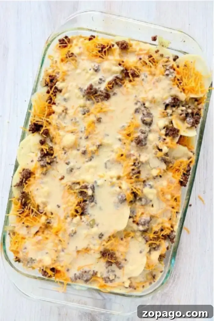 Hamburger Casserole 6 A glass baking dish filled with a delicious Hamburger HotDish showcases its layered casserole. Visible ingredients include sliced potatoes, browned ground beef, melted cheese, and a creamy sauce. The dish rests on a light-colored wooden surface, inviting you to savor its flavors.