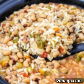 A crockpot filled with black-eyed peas, rice, vegetables, and a savory broth, with a spoon for serving.