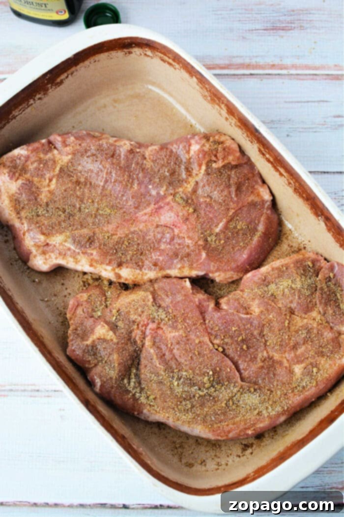 Tender Oven Baked Pork Steaks 5 Two seasoned pork steaks in a ceramic-coated baking dish, with an olive oil bottle and wooden surface in the background, ready for roasting.