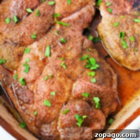 Tender Oven Baked Pork Steaks 7 Close-up of seasoned pork steaks in the oven, perfectly cooked and topped with chopped parsley, nestled in a baking dish.