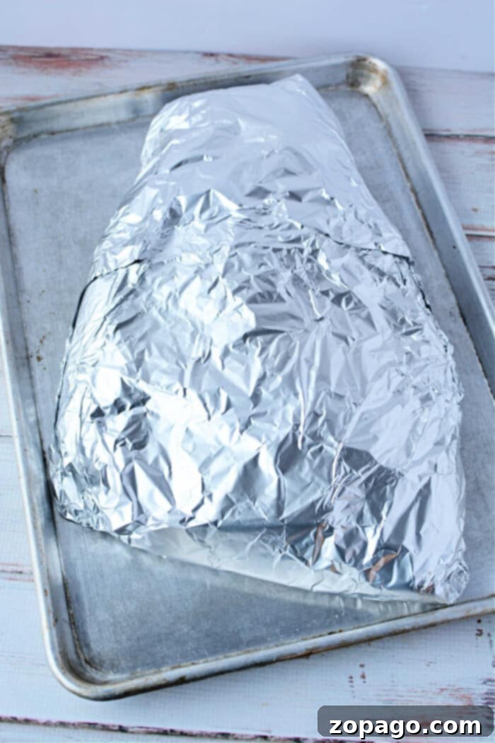 Perfect Shredded Oven Brisket 6 A foil-wrapped brisket in the oven, rests on a baking sheet placed on a white wooden surface.