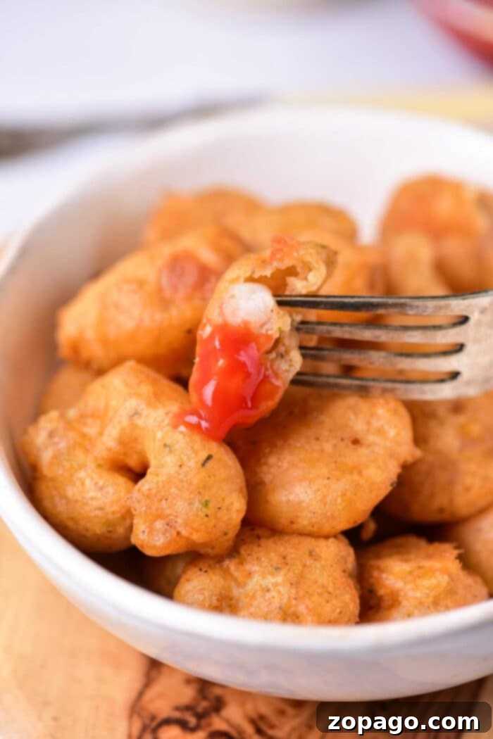Golden Crispy Beer Shrimp 7 A fork holds a beer-battered fried shrimp bite with ketchup in a white bowl of similar bites.