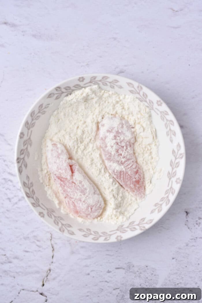 Brewery-Style Crispy Chicken Strips 5 Two raw chicken tenders, are coated in flour and elegantly arranged in a decorative bowl on a light textured surface.