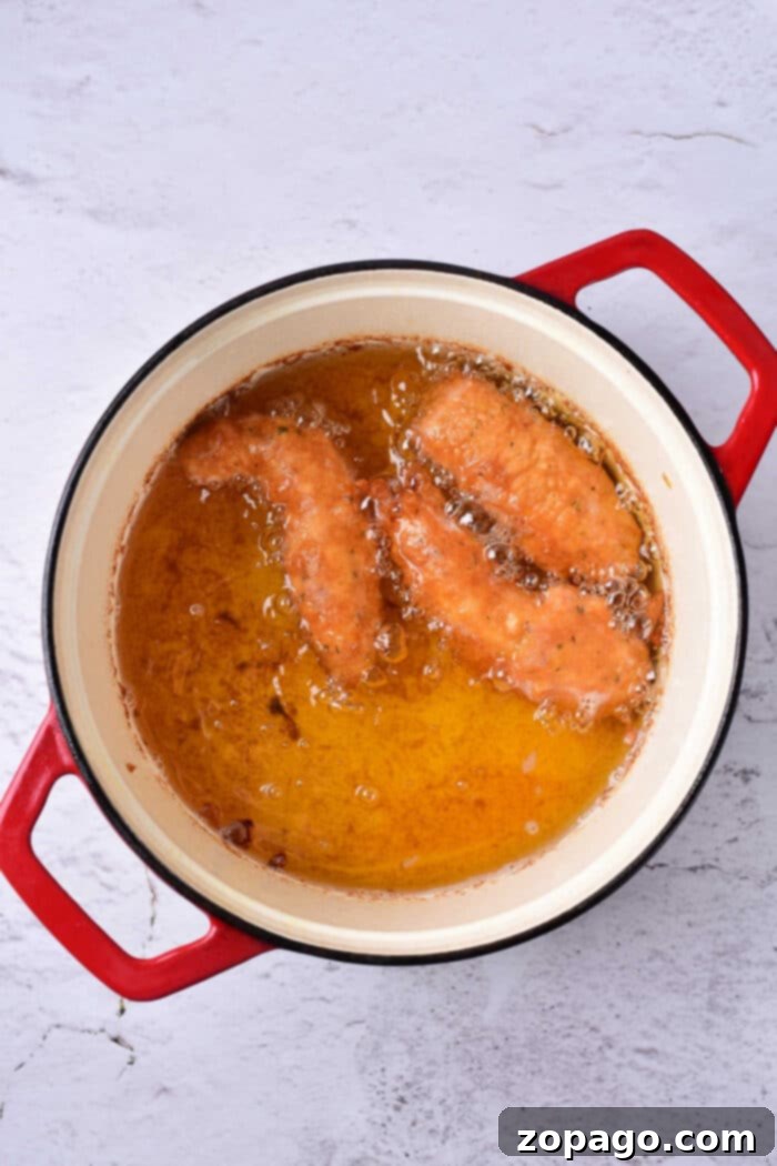 Brewery-Style Crispy Chicken Strips 8 Two beer battered chicken tenders frying in hot oil alongside in a red-handled pot on a light gray surface.