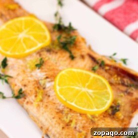 Oven-Baked Trout Perfection 5 baked trout fillet garnished with lemon slices and thyme sits elegantly on a white plate, reminiscent of a dish youd find alongside baked trout. Nearby, a red-striped napkin adds a touch of charm.