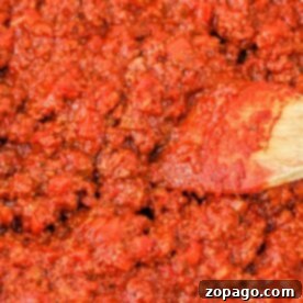 Close-up of a rich, chunky tomato sauce with cherry tomatoes, as a wooden spoon stirs the vibrant red mixture.