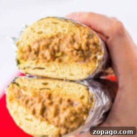 The Chopped Melt 6 A hand holds a chopped cheese sandwich wrapped in foil, its creamy filling spilled slightly as its cut in half.