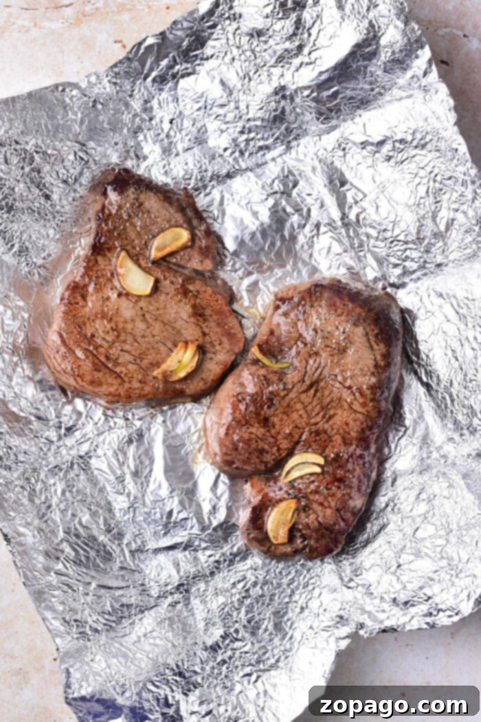 Perfectly Seared Beef Tenderloin Medallions 4 Two succulent cooked beef tenderloin medallions, resting on aluminum foil adorned with delicate garlic slices and fresh herbs for aroma.