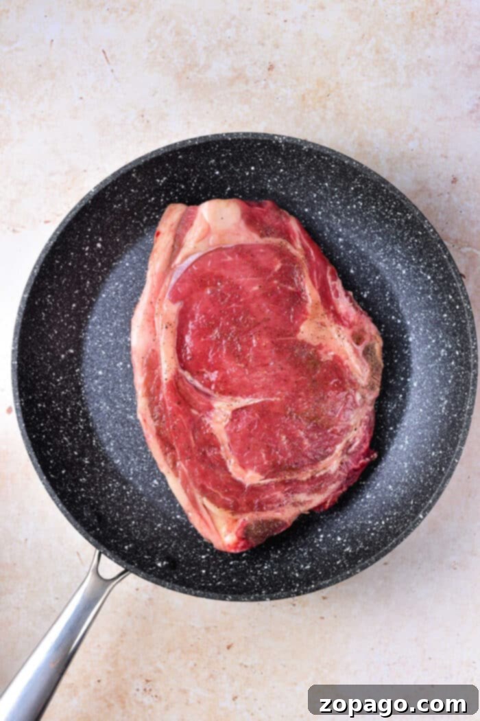 New York Strip Steak 3 Raw New York strip steak, generously seasoned, resting in a preheated black cast iron frying pan, ready for searing as the initial step in oven cooking.