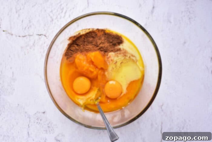 Soulful Southern Sweet Potato Pie 5 A vibrant assortment of sweet potato pie ingredients gathered in a clear glass bowl, resting on a white marbled surface. Mashed sweet potatoes, creamy condensed milk, raw eggs, and an aromatic blend of spices like cinnamon and nutmeg are ready to be mixed. A spoon is placed in the bowl, anticipating the stirring process.
