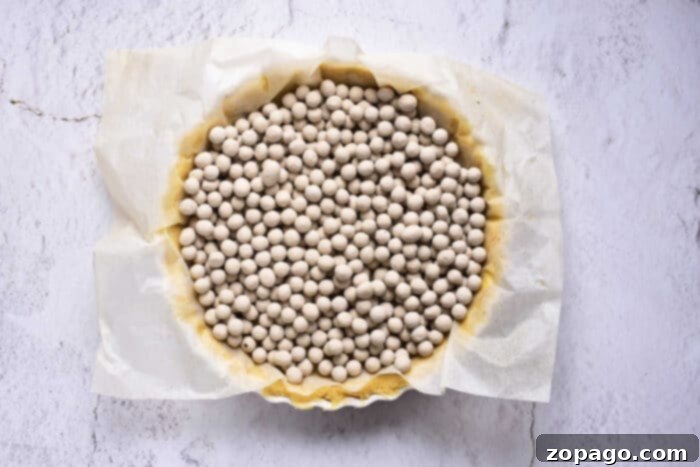Classic Southern Sweet Potato Pie 6 A circular pie crust, meticulously covered with parchment paper and filled with numerous small ceramic baking beads, sits on a pristine white surface. This setup prepares the foundation for a delectable sweet potato pie with sweetened condensed milk, ensuring a perfectly par-baked crust.