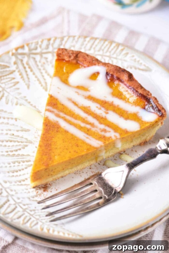 Soulful Southern Sweet Potato Pie 7 A beautifully presented slice of pie, topped with a delicate drizzle of white icing, sits on a decorative white plate. A silver fork rests beside the slice, inviting a taste of this comforting dessert, reminiscent of a homemade sweet potato pie with sweetened condensed milk. The plate is placed on a textured cloth, enhancing its rustic charm.