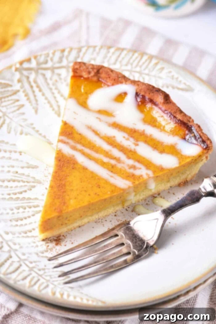 Classic Southern Sweet Potato Pie 7 A generous slice of sweet potato pie, elegantly topped with a delicate drizzle of white icing, is showcased on a decorative white plate. A silver fork rests beside the pie slice, indicating it's ready to be enjoyed. The plate is positioned on a textured cloth, enhancing the visual appeal of this comforting dessert.