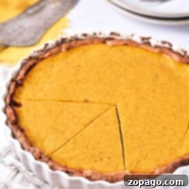A sweet potato pie with a golden-brown crust and smooth filling sits in a white fluted pie dish. A slice has been cut and slightly separated from the rest of the pie. There are plates, forks, and a pie server in the background. A beige cloth is underneath the pie dish.