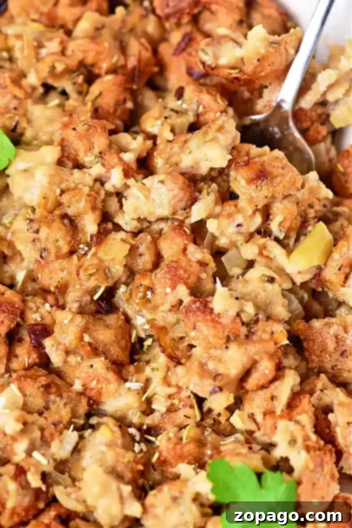Crispy Stuffing, Baked Separately 2 Close-up of a dish of oven-cooked stuffing, showcasing browned bread cubes mixed with cooked onions and herbs. The texture appears moist and chunky, with a spoon partially visible in the top right corner. Garnished with a small parsley leaf.