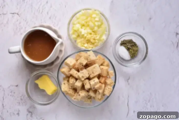 Perfect Pan-Baked Stuffing 3 Ingredients for how to cook stuffing in the oven are laid out on a white textured surface: a pitcher of brown liquid, diced onions and garlic in a small bowl, dried herbs with salt in another, croutons in a medium bowl, and a pat of butter resting on a small plate.