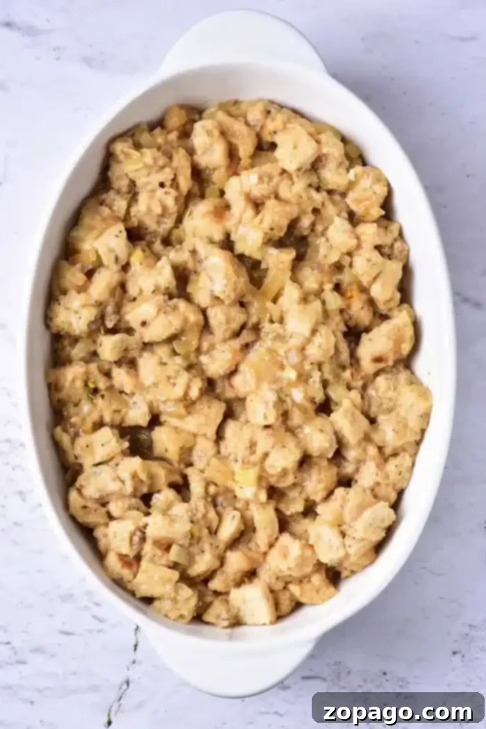 Perfect Pan-Baked Stuffing 5 A white oval dish filled with bread stuffing, containing pieces of bread mixed with herbs and onions, rests on a light-colored marble surface, illustrating how to cook stuffing in the oven.
