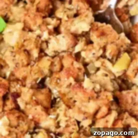Crispy Stuffing, Baked Separately 6 Close-up of a dish of oven-cooked stuffing, showcasing browned bread cubes mixed with cooked onions and herbs. The texture appears moist and chunky, with a spoon partially visible in the top right corner. Garnished with a small parsley leaf.