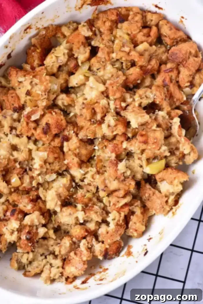 Perfect Pan-Baked Stuffing 7 A close-up of a casserole dish filled with baked stuffing showcases the art of how to cook stuffing in the oven. The golden brown top reveals pieces of bread, herbs, and bits of sautéed onions and celery. A silver serving spoon is partially visible on the right side, ready to serve.