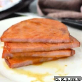 Three slices of ham steak, perfectly glazed and cooked in the oven, are stacked on a white plate with sauce drips.