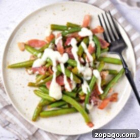 Plate of crack green beans with bacon and creamy sauce, served with a black fork on a light tablecloth.