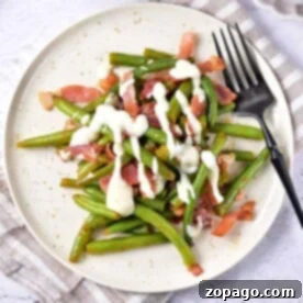 Plate of crack green beans with bacon and creamy sauce, served with a black fork on a light tablecloth.