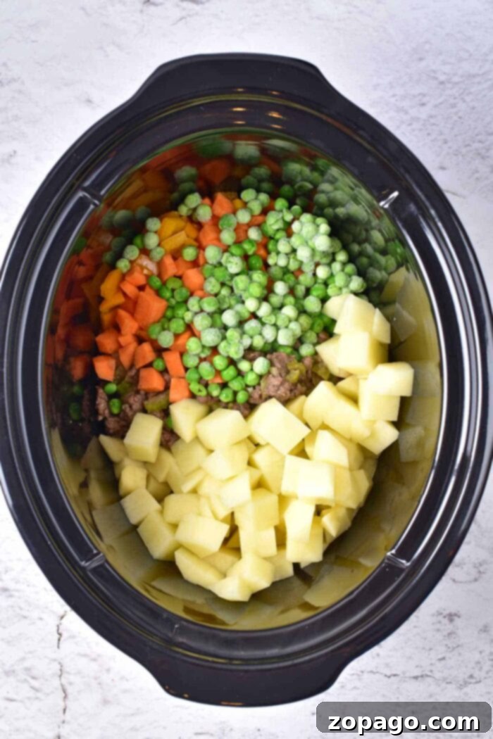 Slow Cooker Creamy Ground Beef and Potato Soup 4 A crockpot brimming with diced potatoes, carrots, peas, and sliced onions sits on a light gray surface, ready to transform into a creamy potato and hamburger soup.