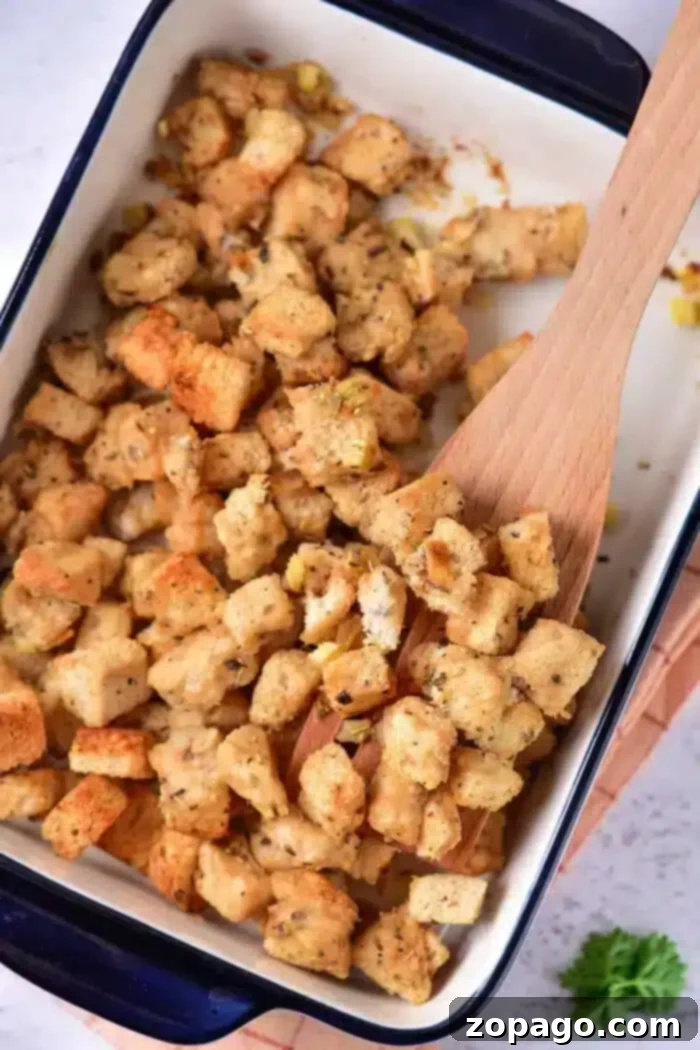 Crispy Sourdough Croutons 2 A baking dish filled with evenly browned sourdough croutons, seasoned with herbs and spices. A wooden spoon rests among the croutons, while a sprig of parsley peeks from the bottom right corner outside the dish.