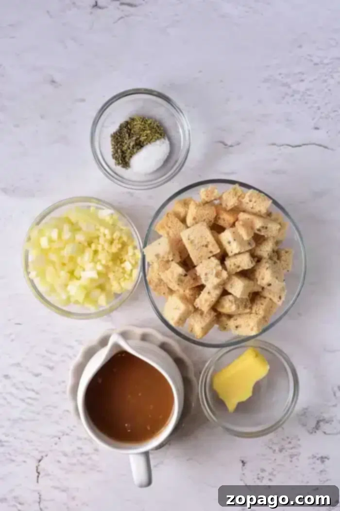 Crispy Sourdough Croutons 3 Top-down view of various ingredients for a stuffing recipe laid out on a light textured surface, including cubed sourdough bread, chopped onions, minced garlic, a small dish of melted butter, a bowl of seasoning with salt and pepper, and a gravy boat filled with brown liquid.