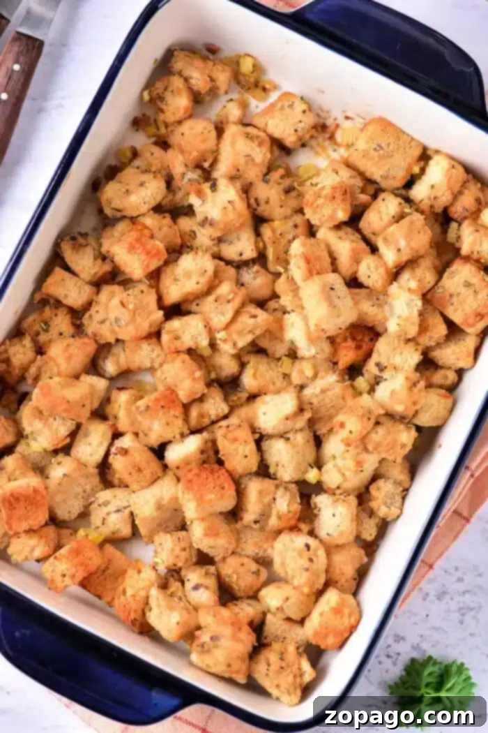 Crispy Sourdough Croutons 7 A baking dish filled with seasoned sourdough croutons, possibly stuffing, arranged evenly. The dish is placed on a light-colored surface with a glimpse of a knife and a green herb nearby.