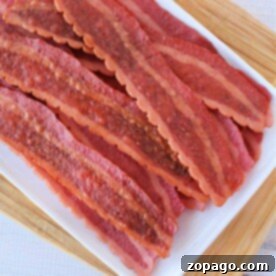 Slices of turkey bacon in the oven are arranged on a white tray atop a wooden board.