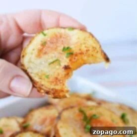 A close-up captures a hand holding a partially eaten crispy potato slice, topped with herbs, above a bowl of oven-roasted sliced potatoes.