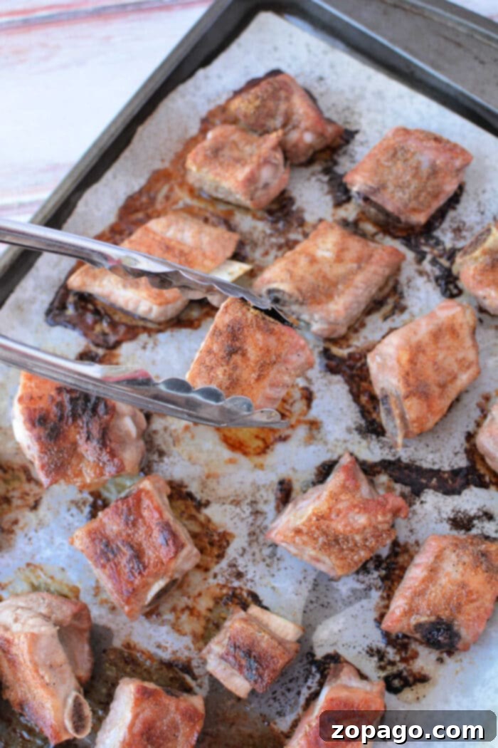 Tender Oven BBQ Riblets 6 Tongs holding a piece of seasoned, baked pork riblet on a tray with multiple baked riblets in the oven.