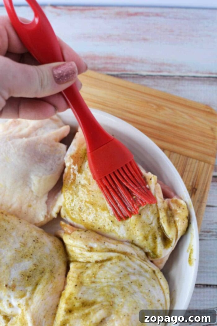 Golden Crispy Chicken Thighs 7 A hand uses a red brush to spread marinade on raw chicken pieces in a white dish, prepping them before discovering how long to bake chicken thighs at 425 for perfect results.