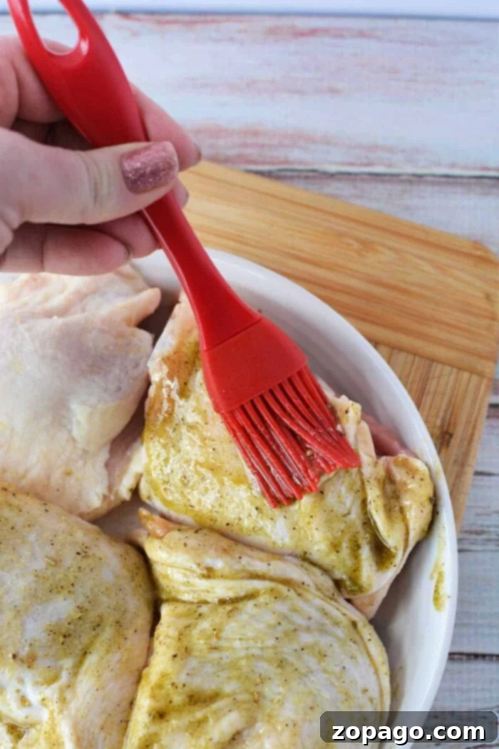 A hand uses a red brush to spread marinade on raw chicken pieces in a white dish, prepping them before discovering how long to bake chicken thighs at 425 for perfect results.