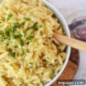 How to Cook Orzo Perfectly 7 A bowl of perfectly cooked orzo pasta garnished with fresh parsley rests on a wooden board, accompanied by a rustic wooden spoon. Curious about crafting this dish? Discover how to cook orzo effortlessly to enjoy at home.